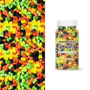 May include: A clear plastic jar filled with colorful bone-shaped sprinkles. The sprinkles are in shades of green, yellow, orange, and black. The jar has a white lid and a label that reads "SugarMeLicious Sprinkles" with the tagline "Sprinkle, Sparkle, Smile."