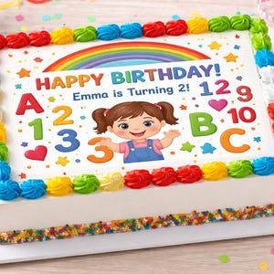 May include: A rectangular birthday cake with white frosting, decorated with colorful frosting borders and sprinkles. The cake features a rainbow, stars, and a cartoon girl. Text on the cake reads "HAPPY BIRTHDAY! Emma is Turning 2!" with numbers and letters.