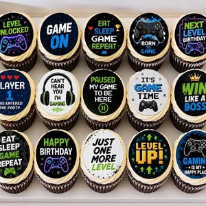 May include: A set of fifteen chocolate cupcakes with white frosting, each topped with a circular edible topper featuring various gaming-themed graphics and phrases like Level Unlocked, Game On, Eat Sleep Game Repeat, and Player 1 Has Entered.