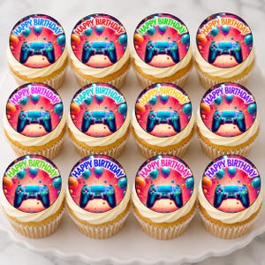 May include: Twelve cupcakes with white frosting, each topped with a circular image. The images feature a video game controller and the words "Happy Birthday" surrounded by balloons. The cupcakes are arranged on a white surface.