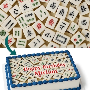 May include: A white cake with blue frosting trim, decorated with edible Mahjong tiles. The cake is topped with the words "Happy Birthday Miriam" in red. The tiles are cream-colored with black, red, and green symbols.