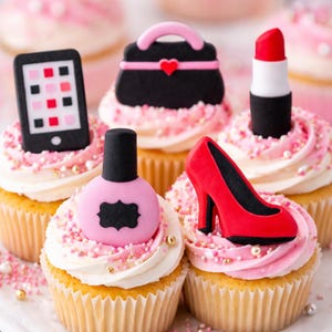 May include: Cupcakes decorated with pink frosting and various themed toppers. Toppers include a black purse, red lipstick, a red high heel, a pink nail polish bottle, and a smartphone. The cupcakes are decorated with pink sprinkles.