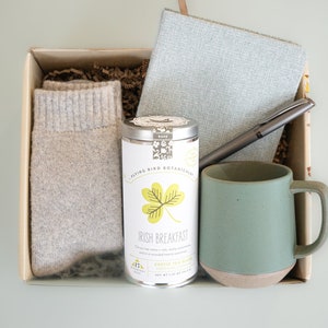 May include: A gift box filled with items for a cozy night in, including a mug, socks, a notebook, a pen, and a tin of Irish Breakfast tea.