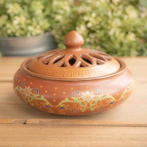 May include: A brown ceramic incense burner with a lid featuring a decorative pattern and a small knob. The body of the burner is adorned with floral and scroll designs in green, blue, and gold. The burner sits on a wooden surface.