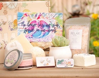 May include: A gift box with various self-care items. Includes a card with text, a succulent in a white pot, bath salts, a candle, a balm tin, a bar of soap, and other small items. The card reads "No longer by our side, forever in our hearts."
