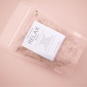 May include: A clear plastic pouch filled with pink bath salts. The pouch has a white label with the text "PURE CHAKRA RELAX Bath Salts Himalayan Pink Salt, Dead Sea Salt, Epsom Salt, Lavender Flowers, Chamomile Flowers, Sweet Orange Essential Oil NET WT 7 oz (200g)"