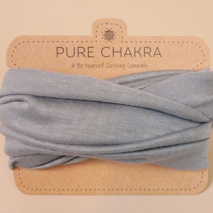 May include: A light blue headband with the text "PURE CHAKRA" on a brown cardboard tag. The tag also includes the text "A Be Yourself Clothing Company" and a sun graphic. The headband is made of a soft, woven material.