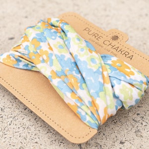 May include: A white fabric headband with a floral pattern in shades of blue, yellow, and green. The headband is packaged on a brown cardboard card with the text "PURE CHAKRA Live. Hope. Live. Free."