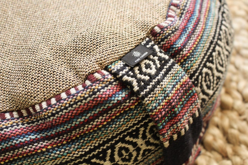 May include: Close-up of a round meditation cushion with a woven fabric top and a colorful patterned fabric bottom. The cushion has a black label with the text "Pure Creation" on it.