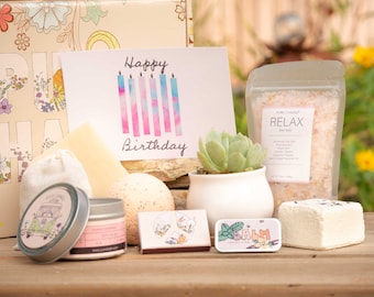 May include: A gift box filled with bath and body products for a birthday celebration. The box includes a card that says "Happy Birthday", a bath bomb, bath salts, a candle, a small succulent plant, and a soap bar.