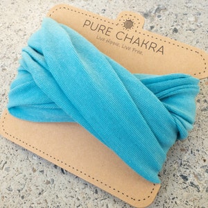 May include: A turquoise headband is displayed on a brown cardboard tag. The tag has the words "PURE CHAKRA" and "Live Hippie. Live Free." printed on it. The headband is made of a soft, textured fabric.