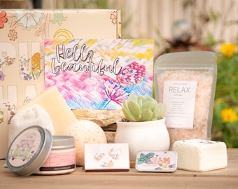 May include: A gift box filled with self-care items, including bath salts, a bath bomb, a candle, soap, and a small succulent plant. The box is decorated with a floral pattern and the words "Hello beautiful".