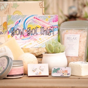 May include: A gift box filled with self-care items, including bath salts, a bath bomb, a candle, a small succulent plant in a white pot, and a card that says "you got this!" The box is decorated with a floral pattern and the word "Pura Vida" in large letters.