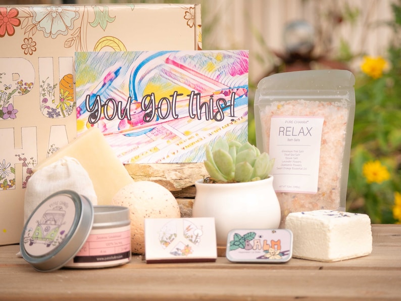 May include: A gift box filled with self-care items, including bath salts, a bath bomb, a candle, a small succulent plant in a white pot, and a card that says "you got this!" The box is decorated with a floral pattern and the word "Pura Vida" in large letters.