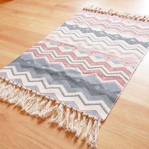 May include: A white, gray, and pink patterned rug with a fringe border. The rug has a chevron pattern.