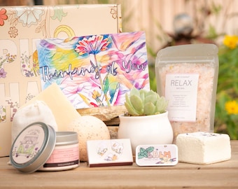 May include: A gift box filled with self-care items, including bath salts, a bath bomb, a candle, and a small succulent plant. The box also includes a card that says "Thinking of You" and a small wooden sign that says "Balm".