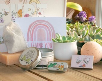 May include: A gift box filled with self-care items, including a pink bath bomb, a small white candle, a tin of lip balm, a small bag of tea, a bar of soap, and a card with a pink rainbow and the text "Thank You".