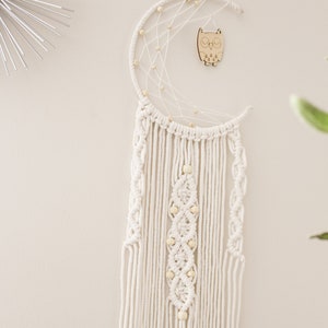 May include: A white macrame dream catcher with a crescent moon shape and wooden owl charm. The dream catcher has white beads and a fringe of white yarn.
