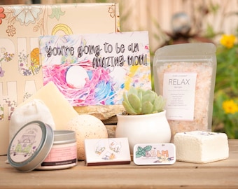 May include: A gift box filled with self-care items for a new mom. The box includes a bath bomb, bath salts, a candle, a small succulent plant, and a card that says "You're going to be an amazing mom!"