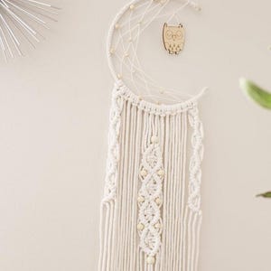 May include: A white macrame dream catcher in a crescent moon shape, adorned with wooden beads and a small wooden owl. The dream catcher features intricate knotting and hanging fringe, creating a bohemian aesthetic. It is designed for wall decor.