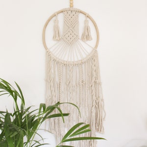 May include: A white macrame dream catcher with a wooden ring and tassels hanging from the bottom. The dream catcher is hanging on a white wall.