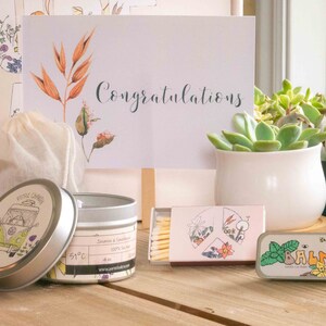 K&ouml;nnte beinhalten: Ein Geschenkset mit einer Kerze, Streichh&ouml;lzern, Lippenbalsam und einer kleinen Tasche. Die Kerze befindet sich in einer silbernen Dose mit einer Illustration eines gr&uuml;nen und gelben Lieferwagens und dem Text "Pure Chill". Die Streichh&ouml;lzer sind in einer rosa und wei&szlig;en Schachtel mit einem Blumendesign. Der Lippenbalsam befindet sich in einer silbernen Dose mit einem gr&uuml;nen Blattdesign und dem Text "BALM". Die Tasche ist wei&szlig; und aus Naturstoff.