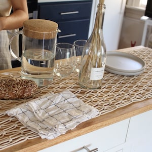 May include: A white crocheted table runner with a woven pattern, a glass pitcher of water, a loaf of bread, a white and blue checkered cloth napkin, and a clear glass bottle with dried flowers and a label that says "Aoccana" on a wooden table.