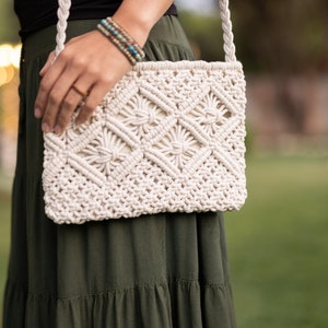 May include: Cream-colored macrame crossbody bag with a geometric pattern. The bag features a woven design and a braided strap. The bag is held by a person wearing a black top and a green skirt. The bag is a stylish accessory.