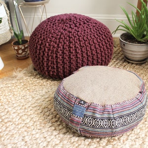 May include: Two round floor cushions, one is a burgundy knitted cushion and the other is a beige cushion with a colorful patterned border. The cushions are on a woven jute rug.