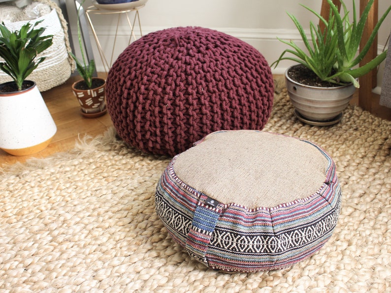 May include: Two round floor cushions, one is a burgundy knitted cushion and the other is a beige cushion with a colorful patterned border. The cushions are on a woven jute rug.