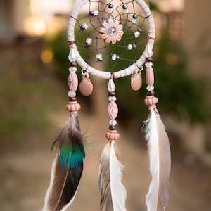 May include: A white dreamcatcher with brown and white feathers, beads, and a pink and white flower design in the center.