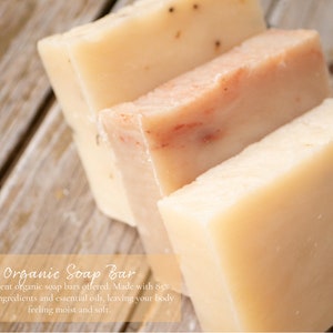 May include: Four bars of organic soap, two are a light beige color and two are a light peach color. The soap bars are stacked on top of each other on a wooden surface. The text "Organic Soap Bar" is written in a light brown font on a light orange background.
