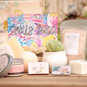 May include: A gift box filled with self-care items, including bath salts, bath bombs, a candle, and a small succulent plant. The box is decorated with a floral pattern and the card reads "Finals are over!"