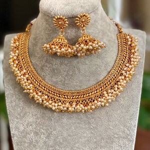 K&ouml;nnte beinhalten: Ein goldenes Halsketten- und Ohrring-Set mit einem filigranen Design und Perlenakzenten. Die Halskette hat ein mehrschichtiges, filigranes Muster mit kleinen Perlentropfen, die von unten h&auml;ngen. Die Ohrringe sind &auml;hnlich gestaltet mit einem kleinen Perlentropfen, der an jedem Ohrring h&auml;ngt.