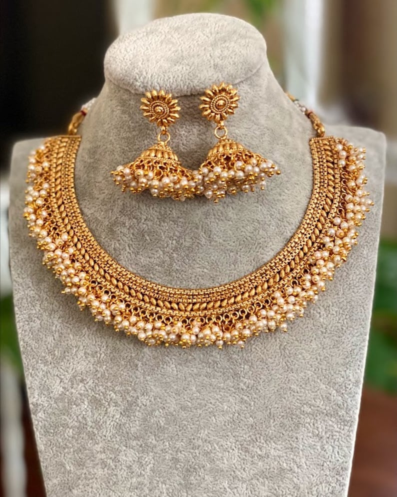 K&ouml;nnte beinhalten: Ein goldenes Halsketten- und Ohrring-Set mit einem filigranen Design und Perlenakzenten. Die Halskette hat ein mehrschichtiges, filigranes Muster mit kleinen Perlentropfen, die von unten h&auml;ngen. Die Ohrringe sind &auml;hnlich gestaltet mit einem kleinen Perlentropfen, der an jedem Ohrring h&auml;ngt.