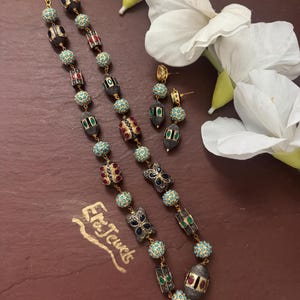 Puede incluir: Un collar largo y pendientes a juego de metal dorado con cuentas de color turquesa y cuentas oscuras texturizadas incrustadas con piedras rojas, verdes y azules. El collar incluye un cierre dorado y el texto Era Jewels es visible.
