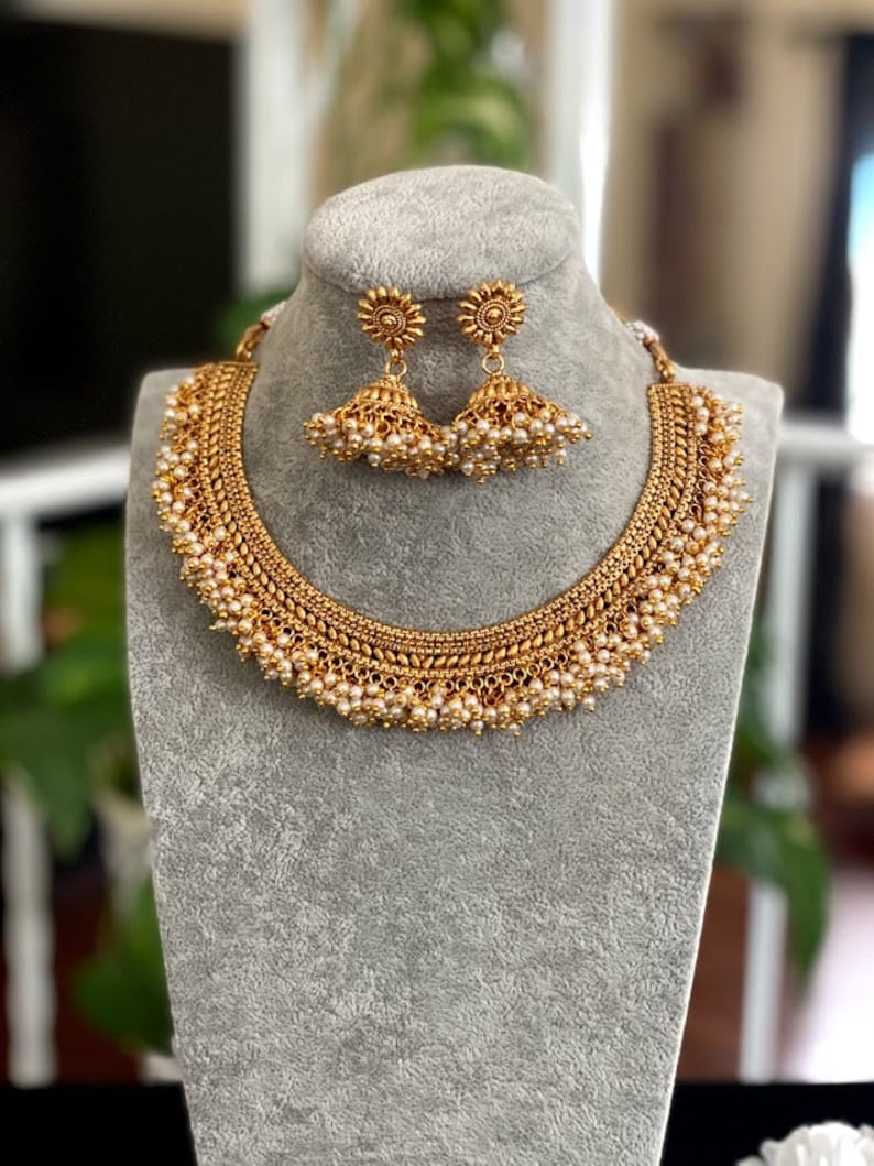 K&ouml;nnte beinhalten: Ein goldenes Halsketten- und Ohrring-Set mit einem zarten Design und Perlenakzenten. Die Halskette hat eine mehrschichtige Kette mit kleinen Perlen, die an jeder Schicht h&auml;ngen. Die Ohrringe sind im H&auml;nge-Stil mit einem Perlen-Tropfen.