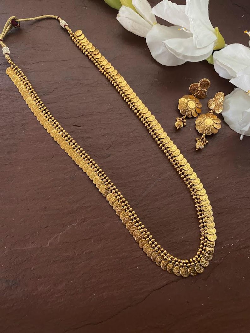 May include: A gold-colored necklace and matching earrings set. The necklace features rows of small, coin-shaped pendants. The earrings have a floral design. The jewelry is displayed on a dark brown surface.