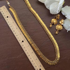 May include: A gold-colored necklace and matching earrings set. The necklace is approximately 28 inches long and features a series of small, coin-shaped pendants. The earrings have a similar design. The jewelry is displayed on a dark brown surface.
