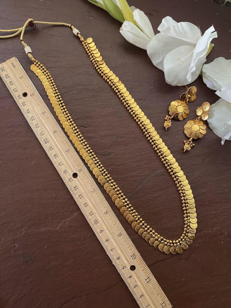 May include: A gold-colored necklace and matching earrings set. The necklace is approximately 28 inches long and features a series of small, coin-shaped pendants. The earrings have a similar design. The jewelry is displayed on a dark brown surface.