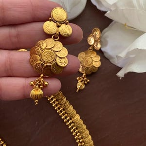 May include: Gold-toned coin earrings and necklace set. The earrings feature a cluster of small, detailed coins, with a dangling beaded accent. The necklace is composed of similar coin-shaped links. The jewelry is displayed against a soft, neutral background.