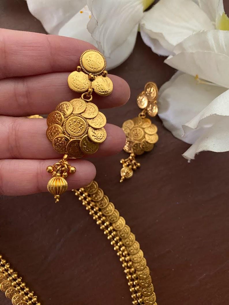 May include: Gold-toned coin earrings and necklace set. The earrings feature a cluster of small, detailed coins, with a dangling beaded accent. The necklace is composed of similar coin-shaped links. The jewelry is displayed against a soft, neutral background.