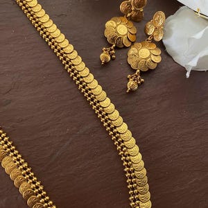 May include: Gold-toned jewelry set featuring a necklace and earrings. The necklace has a coin-like design, and the earrings have a floral motif with dangling accents. The set is displayed on a dark brown surface.