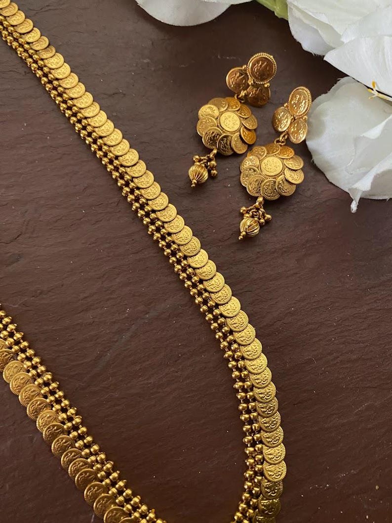 May include: Gold-toned jewelry set featuring a necklace and earrings. The necklace has a coin-like design, and the earrings have a floral motif with dangling accents. The set is displayed on a dark brown surface.