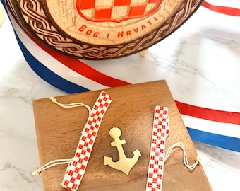 May include: Two red and white checkered bracelets with white string ties, a gold anchor charm, and a wooden box with a gold latch. The box is sitting on a white surface with a red, white, and blue ribbon.