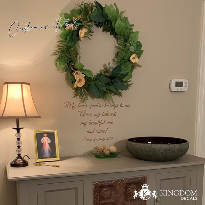 May include: A wall decal with a quote from the Song of Songs: "My lover speaks: he says to me, "Arise, my beloved, my beautiful one, and come!" Song of Songs 2:10". The decal is displayed on a wall with a wreath, a lamp, a picture frame, and a bowl on a table below.