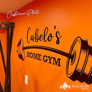 May include: Black and white vinyl decal of a barbell with weights on each end. The decal says "Cabelo's Home Gym" in a cursive font.