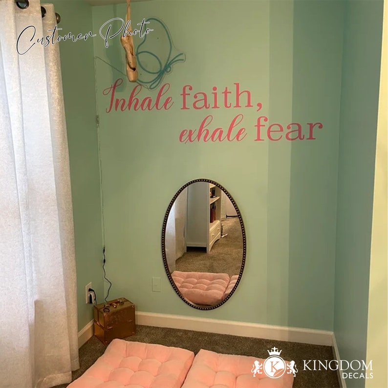 May include: A light green wall with a pink and white wall decal that reads "Inhale faith, exhale fear". A white oval mirror with black trim is on the wall. There are two pink cushions on the floor in front of the mirror.