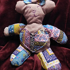 May include: A handmade plush bear with a heart-shaped chest and patterned fabric accents. The bear has a light brown body, embroidered facial features, and a small bow on its head. The colorful fabric includes floral and paisley designs.