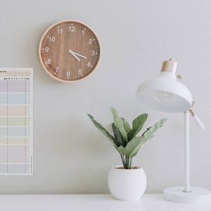 May include: A white desk with a wooden clock, a white lamp, and a potted plant. A printed sheet with the text "Haushaltsplan" is on the wall.